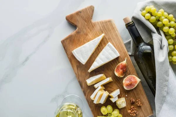 flat lay with food composition of camembert cheese on cutting board, wine and fruits on white marble surface