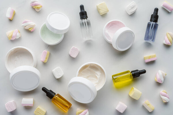 top view of bottles of cream, natural oils and marshmallows on white surface, beauty concept