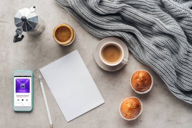 top view of cup of coffee with muffins, blank paper and smartphone with music player app on screen on concrete surface with knitted wool drapery