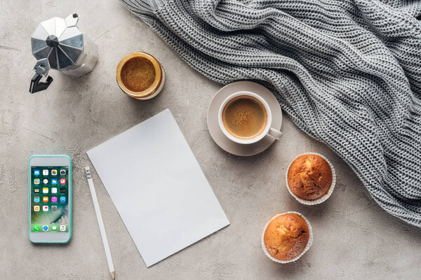 top view of cup of coffee with muffins, blank paper and smartphone with ios homescreen on concrete surface with knitted wool drapery