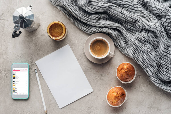 top view of cup of coffee with muffins, blank paper and smartphone with music playlist on screen on concrete surface with knitted wool drapery