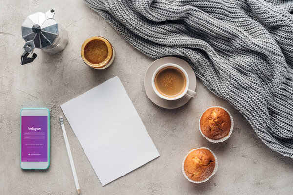 top view of cup of coffee with muffins, blank paper and smartphone with instagram app on screen on concrete surface with knitted wool drapery