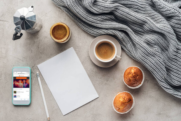 top view of cup of coffee with muffins, blank paper and smartphone with soundcloud app on screen on concrete surface with knitted wool drapery