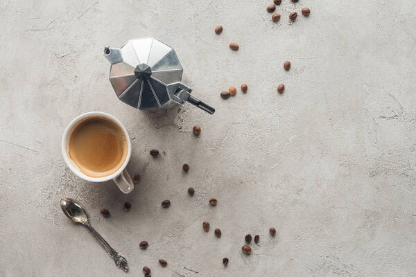 top view of cup of coffee and moka pot on concrete surface with spilled coffee beans