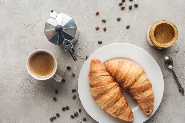 top view of cup of delicious coffee with croissants and moka pot on concrete surface with spilled coffee beans