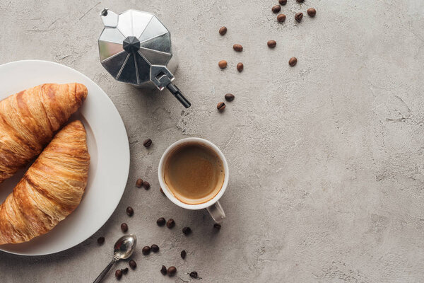top view of cup of coffee with croissants and moka pot on concrete surface with spilled coffee beans