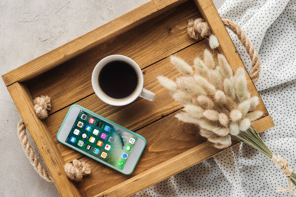 top view of cup of coffee and smartphone with ios homescreen website on screen on tray with lagurus ovatus bouquet on concrete surface