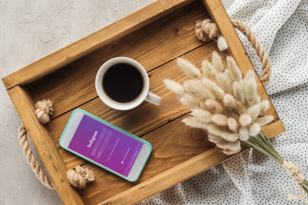 top view of cup of coffee and smartphone with instagram app on screen on tray with lagurus ovatus bouquet on concrete surface