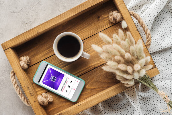top view of cup of coffee and smartphone with music player playing fall out boy on screen on tray with lagurus ovatus bouquet on concrete surface