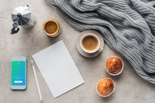 Top view of cup of coffee with muffins, blank paper and smartphone with twitter app on screen on concrete surface with knitted wool drapery — Stock Image Top View Cup Coffee Muffins Blank Paper Smartphone Twitter App Stock Photo