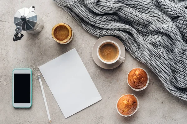 Top view of cup of coffee with muffins, blank paper and smartphone with blank screen on concrete surface with knitted wool drapery — Stock Image Top View Cup Coffee Muffins Blank Paper Smartphone Blank Screen Stock Photo