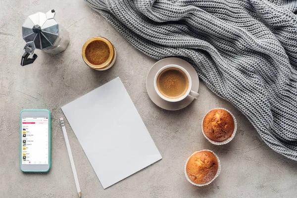 Top view of cup of coffee with muffins, blank paper and smartphone with music playlist on screen on concrete surface with knitted wool drapery — Stock Image Top View Cup Coffee Muffins Blank Paper Smartphone Music Playlist Stock Photo