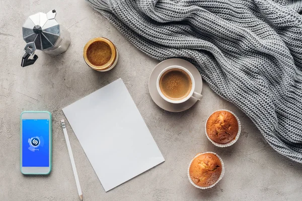 Top view of cup of coffee with muffins, blank paper and smartphone with shazam app on screen on concrete surface with knitted wool drapery — Stock Image Top View Cup Coffee Muffins Blank Paper Smartphone Shazam App Royalty Free Stock Photos