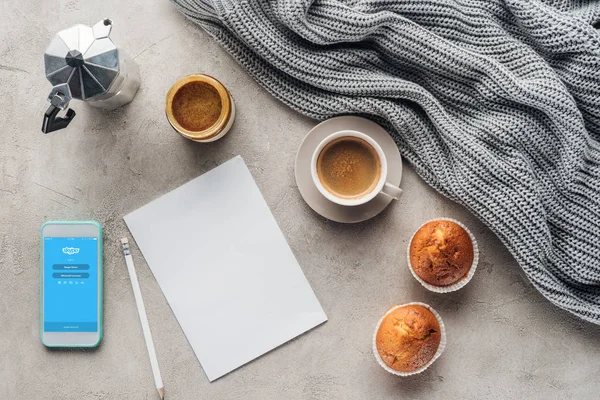 Top view of cup of coffee with muffins, blank paper and smartphone with skype app on screen on concrete surface with knitted wool drapery — Stock Image Top View Cup Coffee Muffins Blank Paper Smartphone Skype App Royalty Free Stock Images