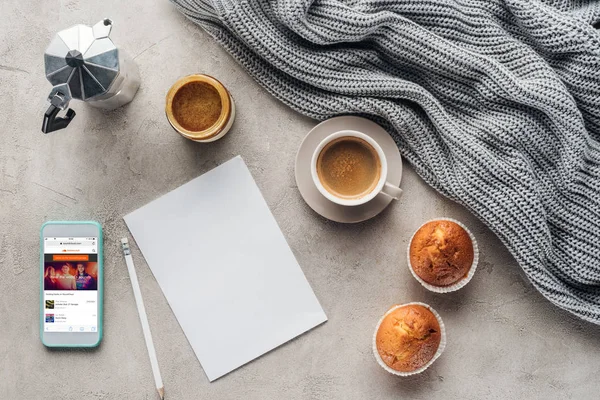 Top view of cup of coffee with muffins, blank paper and smartphone with soundcloud app on screen on concrete surface with knitted wool drapery — Stock Image Top View Cup Coffee Muffins Blank Paper Smartphone Soundcloud App Royalty Free Stock Photos