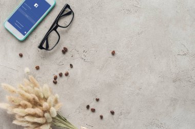 top view of smartphone with facebook app on screen with eyeglasses, spilled coffee beans and lagurus ovatus bouquet on concrete surface clipart