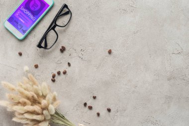 top view of smartphone with shopping app on screen with eyeglasses, spilled coffee beans and lagurus ovatus bouquet on concrete surface clipart