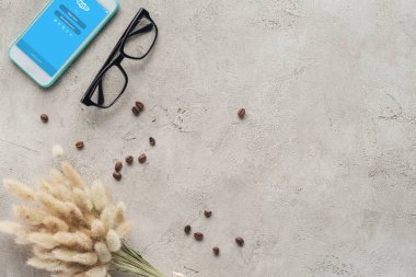 top view of smartphone with skype app on screen with eyeglasses, spilled coffee beans and lagurus ovatus bouquet on concrete surface clipart