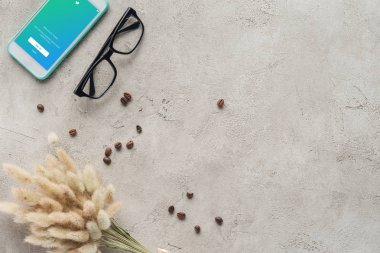 top view of smartphone with twitter app on screen with eyeglasses, spilled coffee beans and lagurus ovatus bouquet on concrete surface clipart