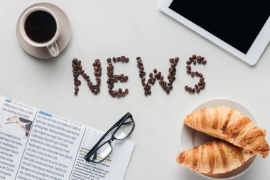kahve kahve çekirdekleri, kruvasan ve beyaz mermer tablete gazeteyle yapılan Haberler yazı ile üstten görünüm