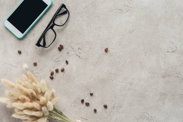top view of smartphone with blank screen with eyeglasses, spilled coffee beans and lagurus ovatus bouquet on concrete surface