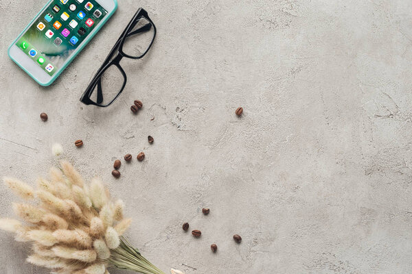 top view of smartphone with ios homescreen with eyeglasses, spilled coffee beans and lagurus ovatus bouquet on concrete surface
