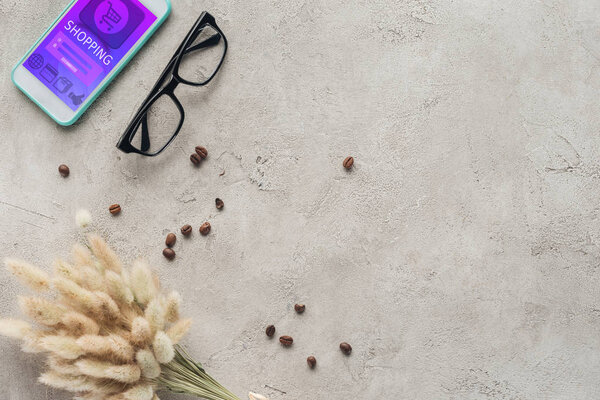 top view of smartphone with shopping app on screen with eyeglasses, spilled coffee beans and lagurus ovatus bouquet on concrete surface