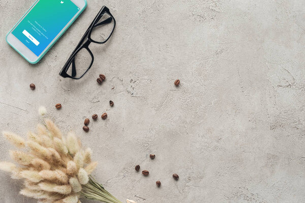 top view of smartphone with twitter app on screen with eyeglasses, spilled coffee beans and lagurus ovatus bouquet on concrete surface