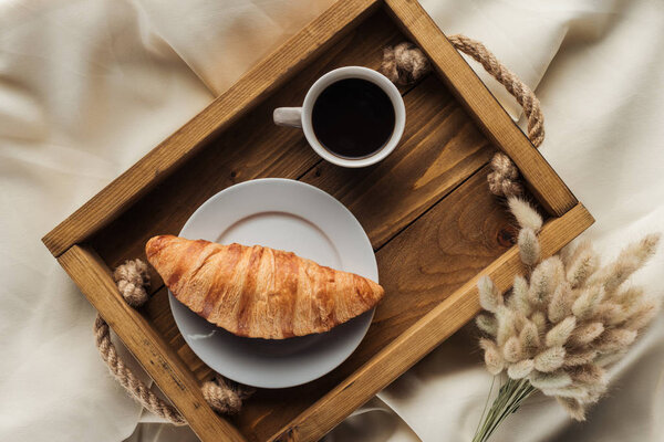top view of coffee and croissant on tray with lagurus ovatus bouquet on beige cloth, breakfast in bed concept