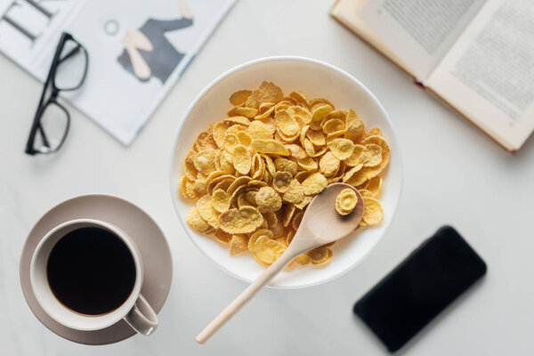 top view of bowl of corn flakes breakfast with cup of coffee and croissant on white surface with newspaper, smartphone and book