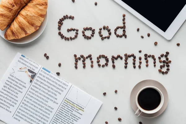 stock image top view of cup of coffee and good morning lettering made of coffee beans on white surface with croissants, newspaper and tablet