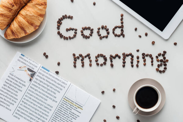 top view of cup of coffee and good morning lettering made of coffee beans on white surface with croissants, newspaper and tablet