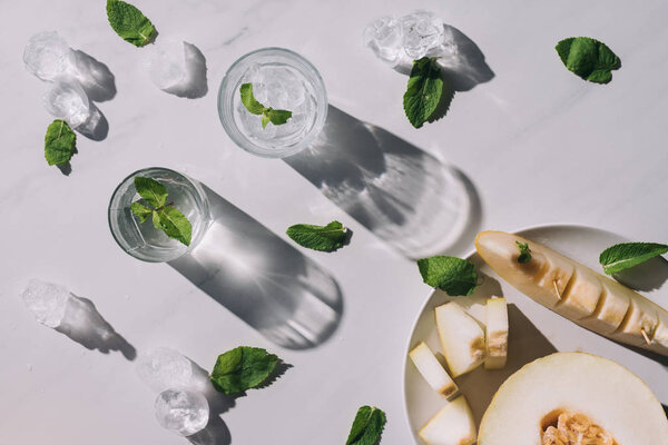 top view of refreshing beverage with mint in glasses and sliced melon with ice cubes on white