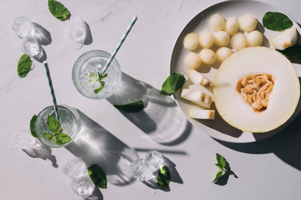 top view of sweet melon balls, half of ripe melon and refreshing beverage with mint and drinking straws in glasses on white 