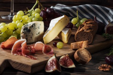 different types of cheeses, prosciutto and grapes on cutting board clipart