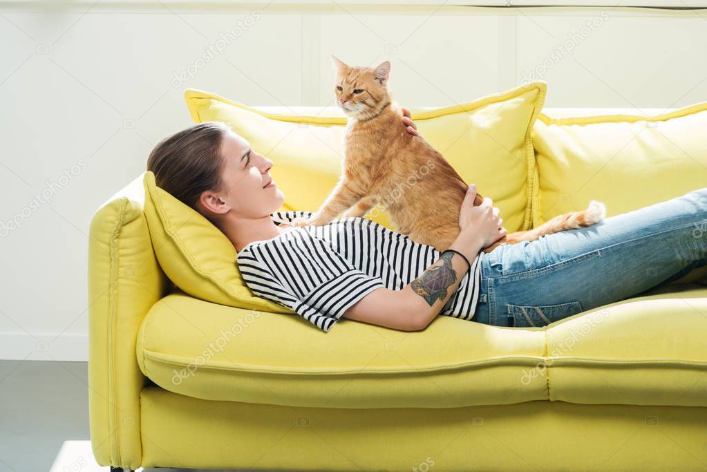 beautiful girl lying on yellow sofa and palming cute red cat in living room