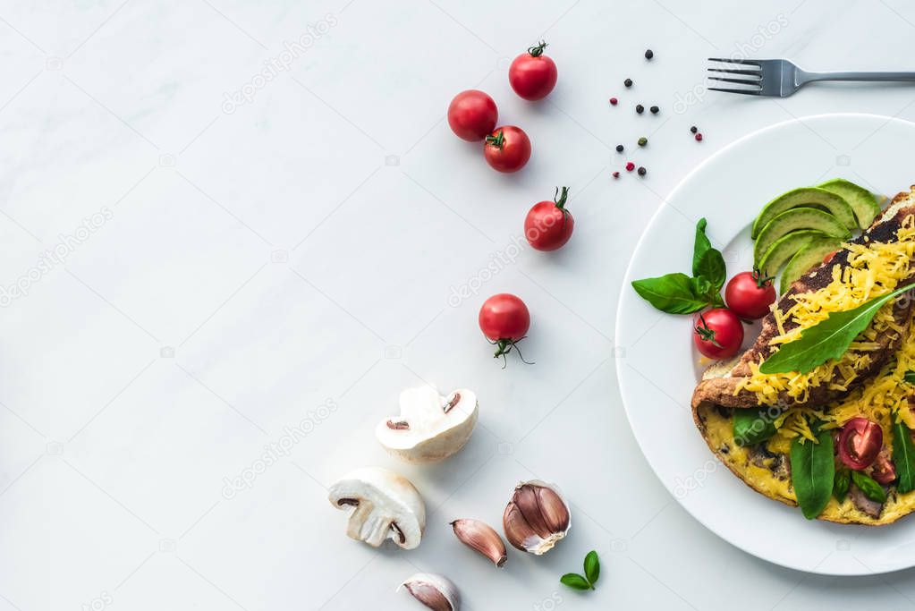 top view of homemade omelette with cherry tomatoes, avocado pieces and cutlery on white marble surface