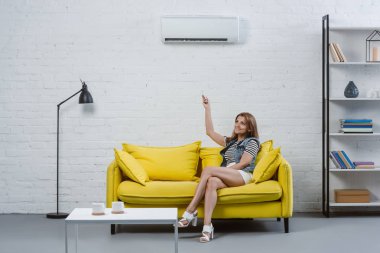 beautiful young woman sitting on sofa and pointing at air conditioner with remote control clipart