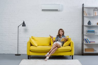 focused young woman with book sitting on sofa and pointing at air conditioner with remote control clipart