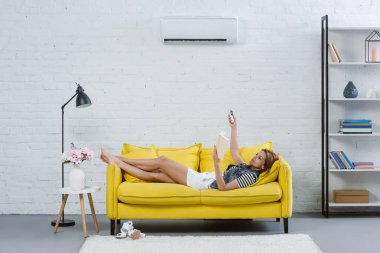 attractive young woman reading book on sofa and pointing at air conditioner with remote control clipart