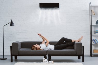 duvarda asılı Klima altında kanepede Smartphone kullanarak rahat genç kadın