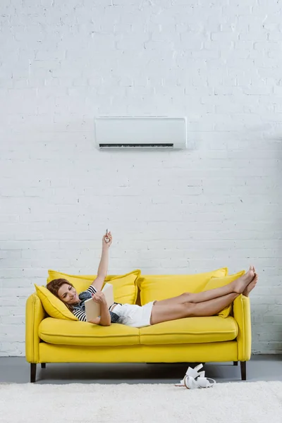 Happy Young Woman Reading Book Sofa Pointing Air Conditioner Remote — Stock Photo, Image