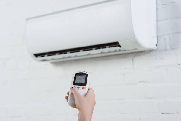 cropped shot of woman pointing at air conditioner hanging on white brick wall with remote control