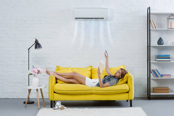 concentrated young woman reading book on couch and pointing at air conditioner hanging on wall with remote control