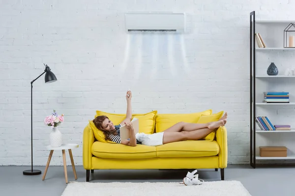 Happy Young Woman Reading Book Couch Pointing Air Conditioner Hanging — Stock Photo, Image