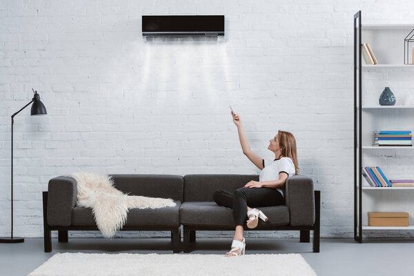 happy young woman pointing at air conditioner hanging on wall with remote control