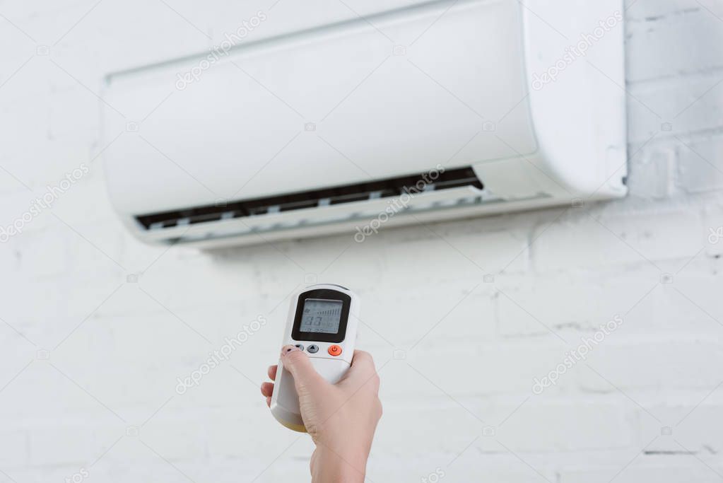 cropped shot of woman pointing at air conditioner hanging on white brick wall with remote control