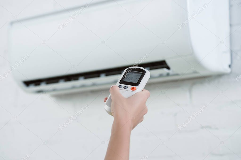 cropped shot of woman pointing at air conditioner hanging on brick wall with remote control