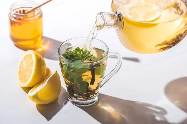 从茶壶倒入柠檬和薄荷的健康茶到杯上白色桌面 — 图库照片