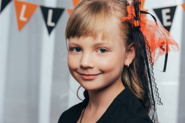 portrait of adorable smiling kid looking at camera wit halloween flags hanging behind at home clipart